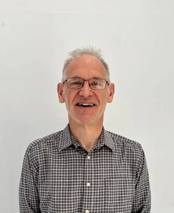 A smiling middle-aged man with glasses and gray hair, wearing a black and white checkered shirt, standing against a plain white wall.