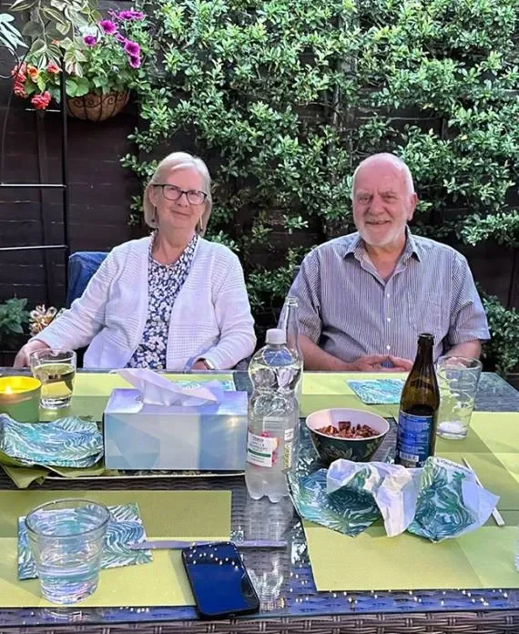 An elderly couple sitting at an outdoor table with food and drinks, surrounded by green plants and flowers.