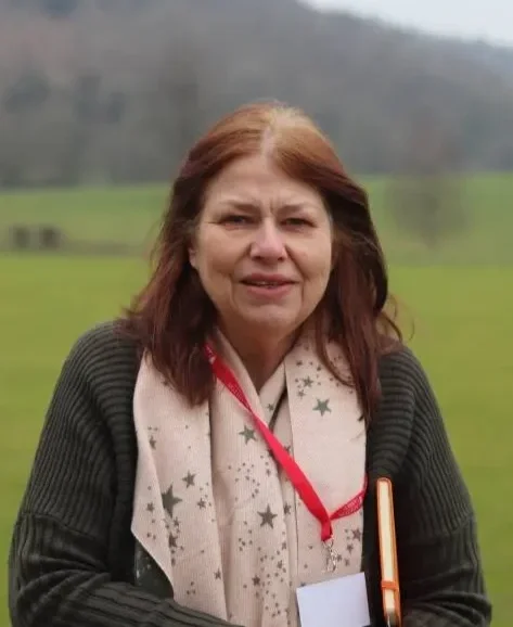 A woman with reddish-brown hair standing outdoors in a green field with a hilly background.