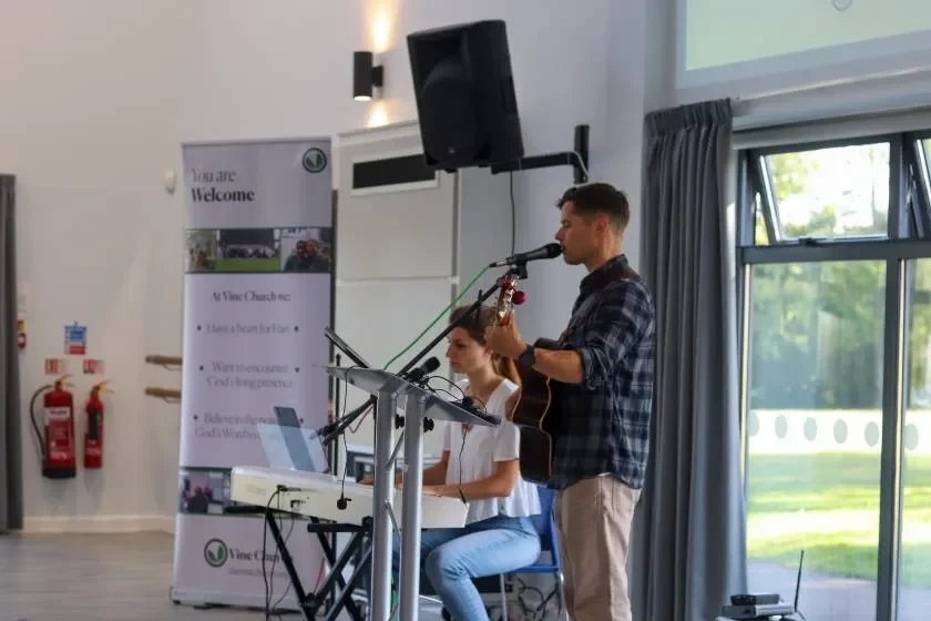 A young man playing guitar and singing into a microphone while a young woman plays keyboard during a performance in a well-lit room with large windows and curtains.