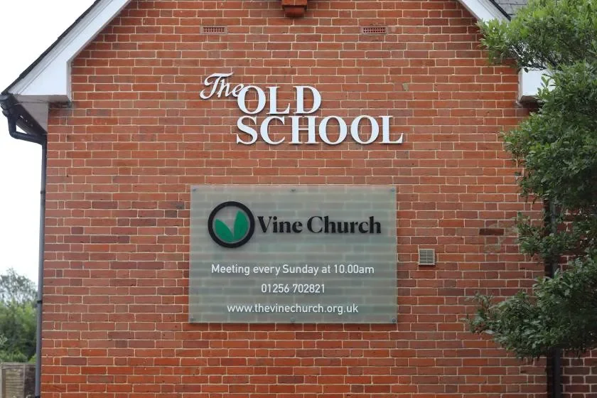 Brick church building with signs indicating it is the old school and home of Vine Church, which meets every Sunday at 10:00 am, with contact and website information.