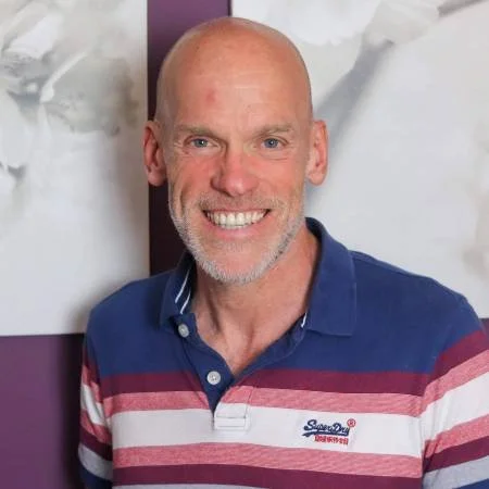 A smiling middle-aged man with a bald head and gray beard, wearing a navy and red striped polo shirt, standing indoors with a white decorative wall behind him.