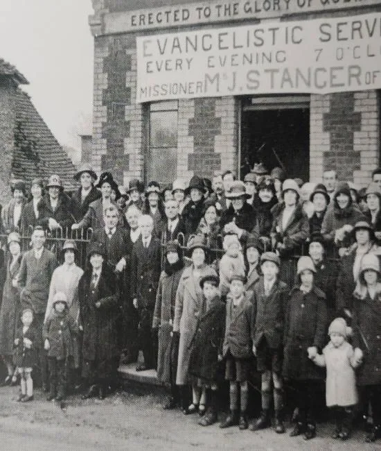 Black and white photo of a large group of people, including children and adults, standing in front of a brick church with a sign that reads "EvangeliSTIC SERVICE EVERY EVENING 7 O'CLOCK MISSIONARY M. J. STANCER."