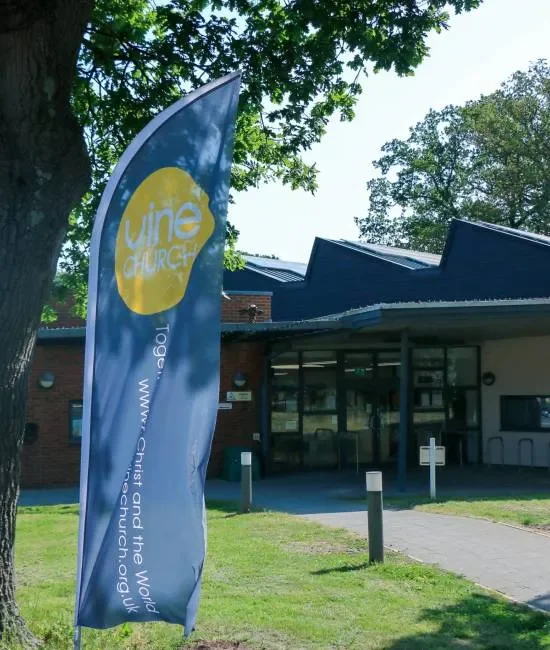 A blue vertical flag with a yellow circle and white text that reads 'Vine Church' and the website URL 'www.church.org', positioned outside a building with a glass entrance, trees, and a grass lawn.