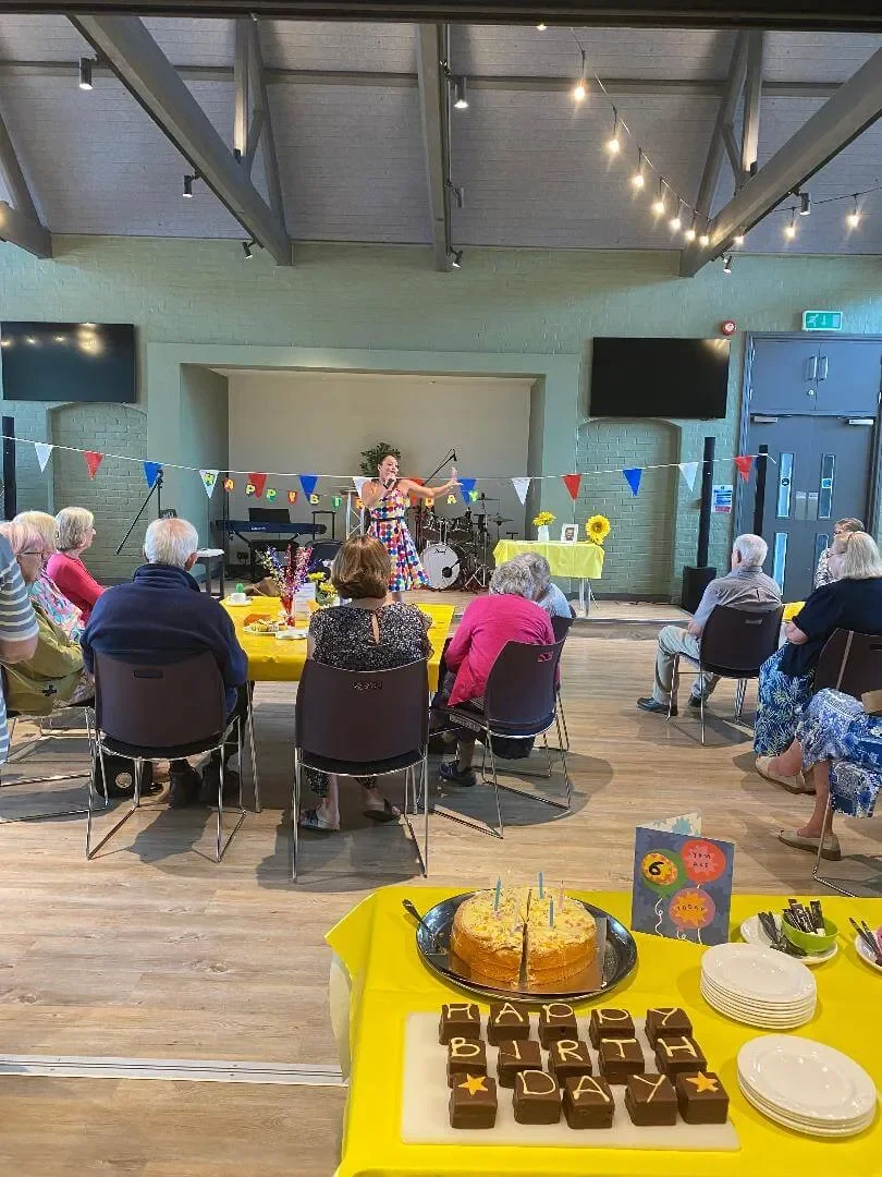 A birthday celebration at a community hall with an elderly group. There is a woman in a colorful dress singing or speaking on stage, with a band in the background. The room is decorated with colorful banners and yellow tablecloths. On the front table, a birthday cake with candles and a tray of chocolate treats spelling out 'HAPPY BIRTHDAY' is visible, along with plates and utensils. The atmosphere appears festive and cheerful.