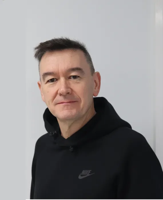 A man with short dark hair and fair skin, wearing a black Nike hoodie, standing against a plain light-colored wall.