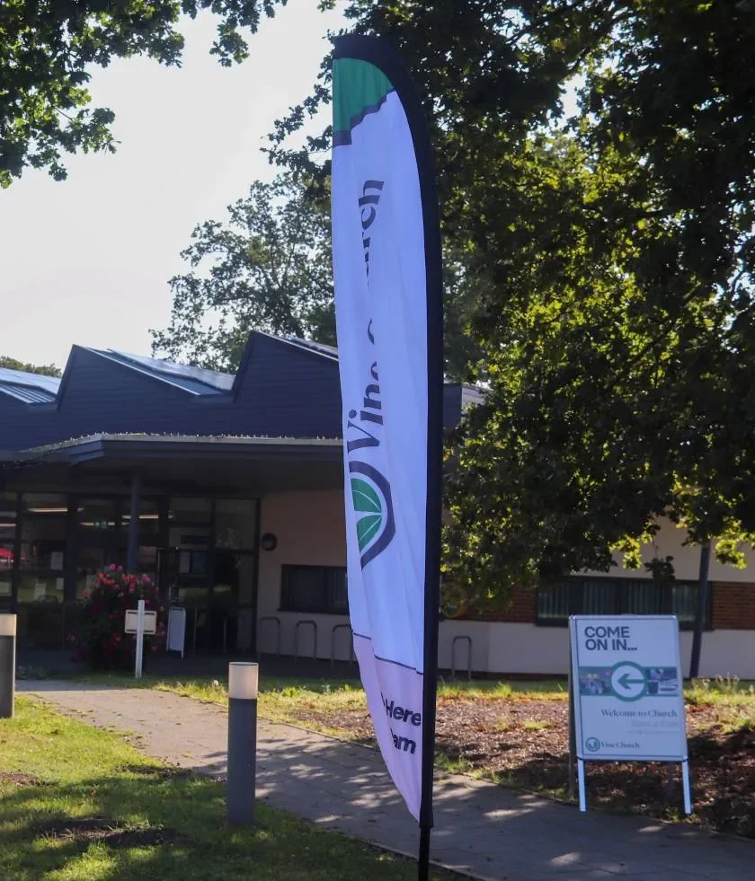 A vertical flag with the word 'Vine' on it, standing outside near a church building. A sign inviting people to enter the church is visible nearby.