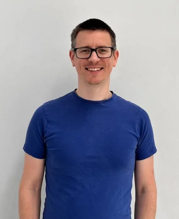 A smiling man with short dark hair, glasses, and a goatee, wearing a blue t-shirt, standing against a plain white background.