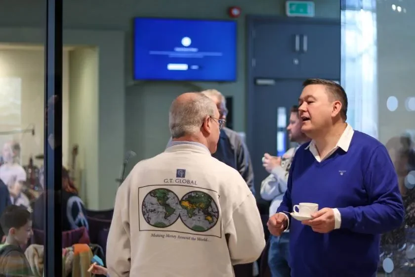 Two men are engaged in conversation through a glass window; the man facing away wears a light-colored jacket with a world map on the back and the words 'Making Almo Americas the World...'; the man facing signifies a tea or coffee cup and saucer in his hand, wearing a blue sweater over a collared shirt; several people are in the background inside a room, some sitting and some standing.