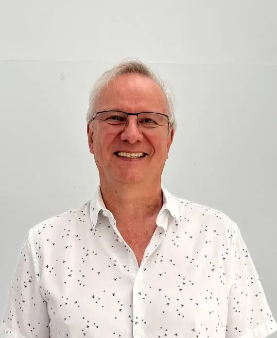 A smiling middle-aged man with glasses, wearing a white button-up shirt with small black star patterns, standing against a plain white wall.
