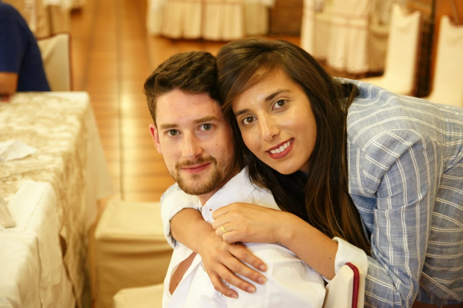 A young man and woman taking a selfie outdoors by a lake with snow-capped mountains in the background, wearing warm jackets and smiling.