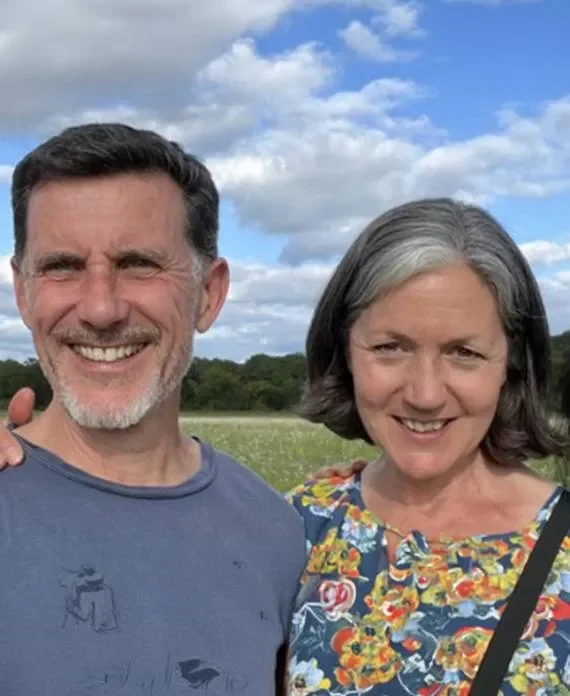 A man and a woman smiling outdoors in a field with green trees and blue sky with clouds behind them.