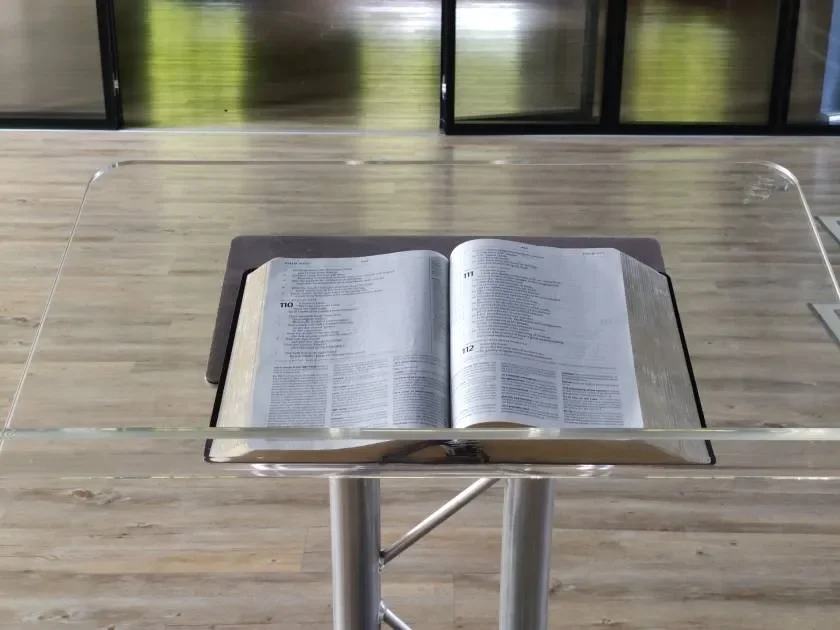 Open Bible on a clear acrylic lectern in a modern, bright room with wooden flooring and large glass windows.