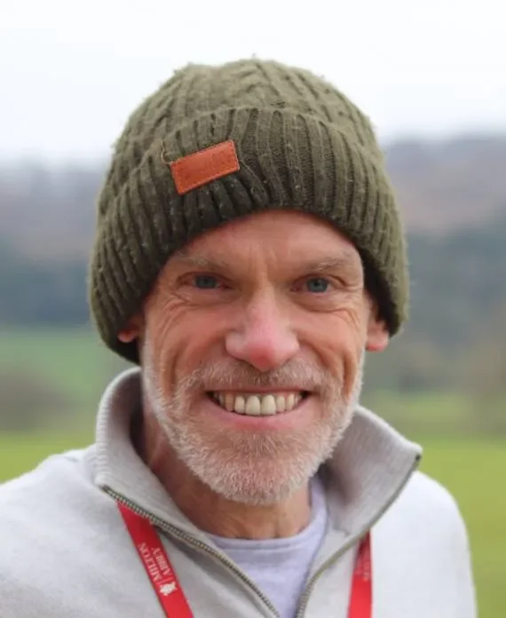 Close-up of a smiling man wearing a green knit beanie and a light gray zip-up jacket outdoors with a blurred green landscape in the background.