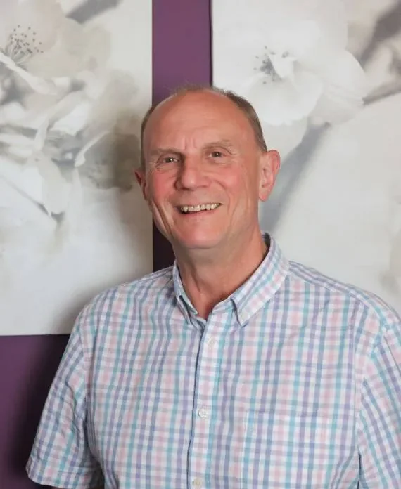 A smiling middle-aged man with a checkered shirt standing in front of white floral artwork on a purple wall.