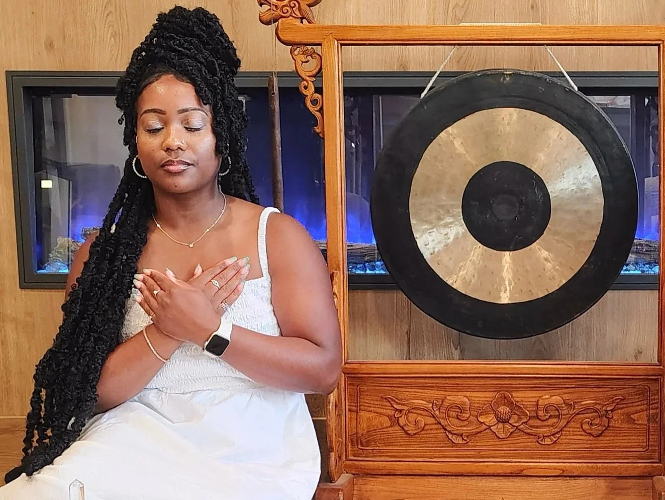 A woman with long black braided hair sitting with her eyes closed, wearing a white dress and jewelry, in front of a large gong attached to a wooden frame, with a fireplace in the background.