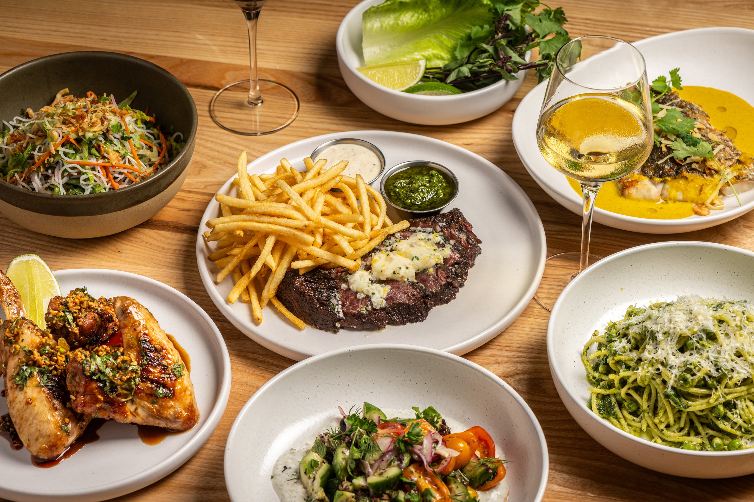 A variety of dishes on a wooden table including a salad, French fries with dipping sauces, grilled meat topped with butter, a vegetable salad, and a fish dish with herbs, lemon, and sauce, accompanied by glasses of white wine.