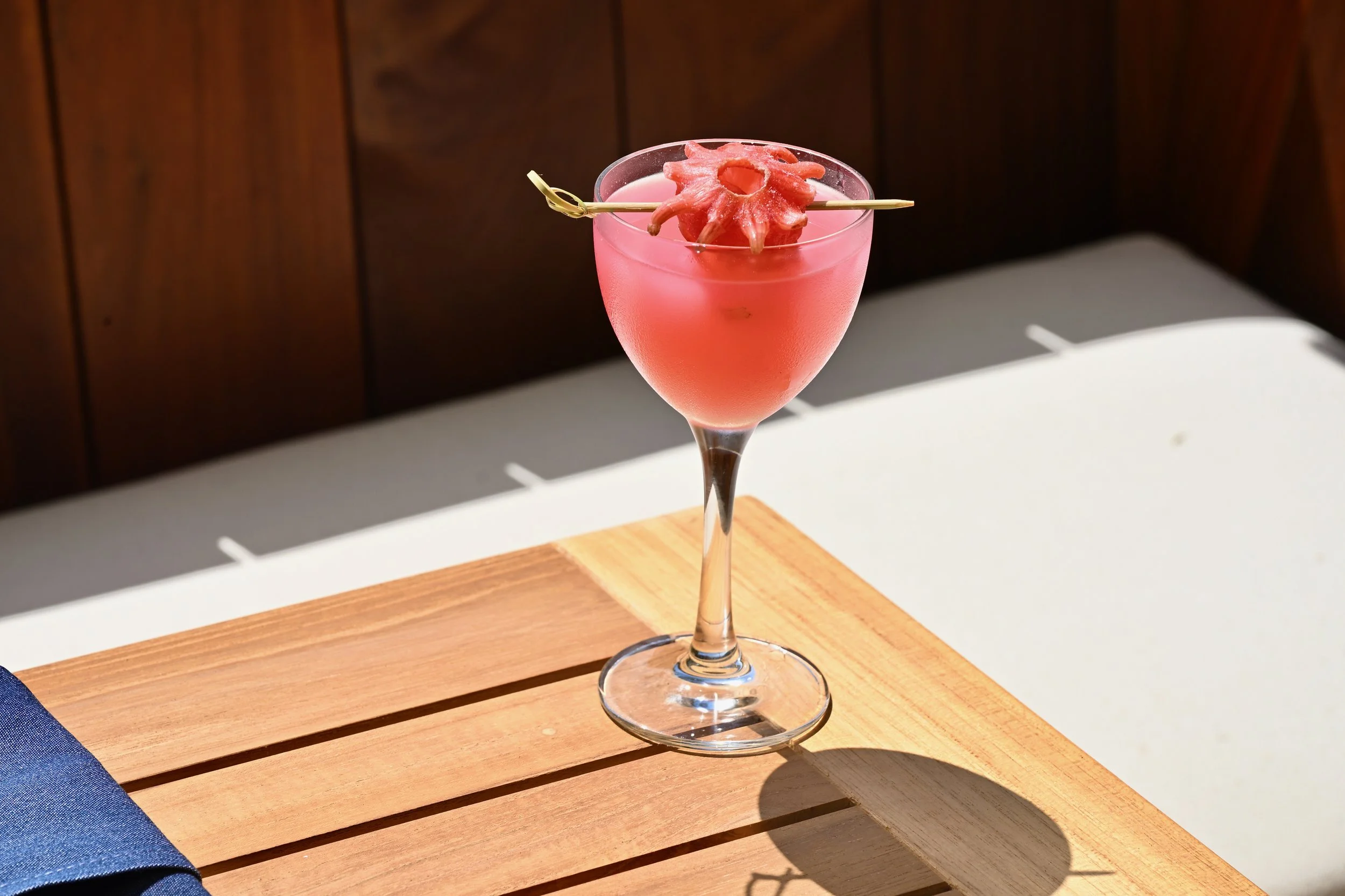 A pink cocktail with a decorative pink flower garnish on a skewer in a tall glass, placed on a wooden surface with sunlight casting shadows.