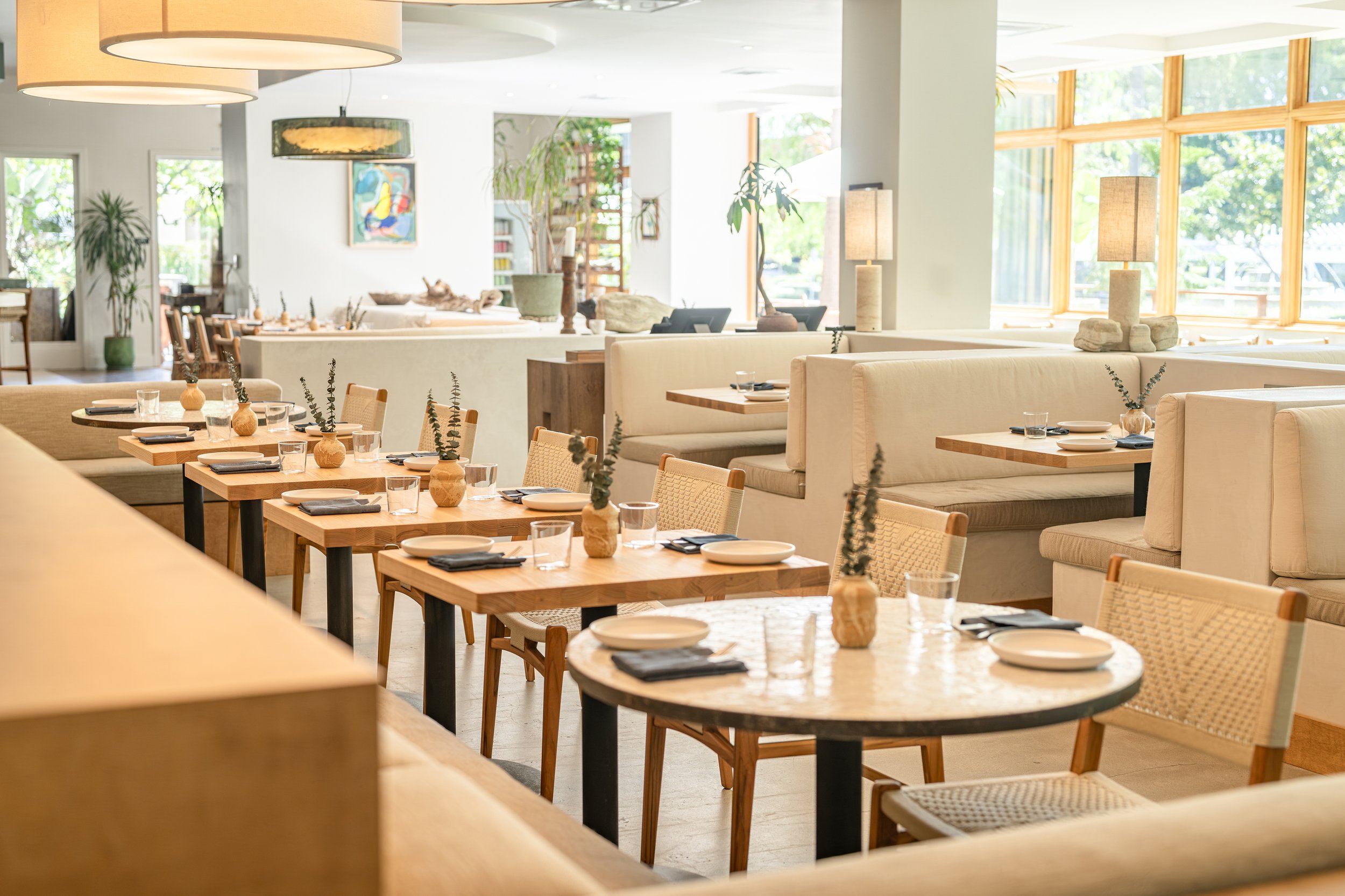 A bright, modern restaurant interior with wooden tables set for dining, including plates, glasses, and napkins. The space features large windows with natural light, beige cushioned chairs, and minimalistic decor with potted plants and abstract artwork on the walls.