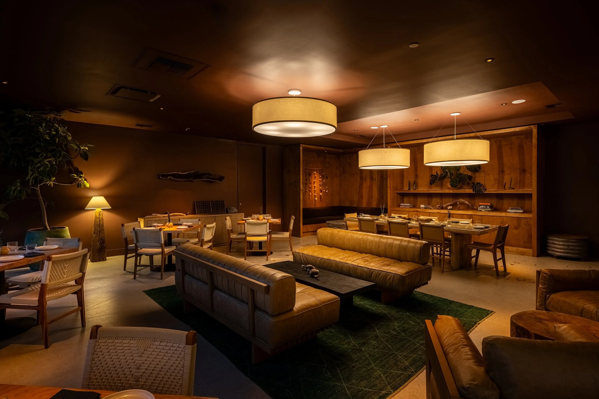 A dimly lit restaurant or lounge with wooden accents and modern pendant lighting, featuring sofas and tables set for dining.