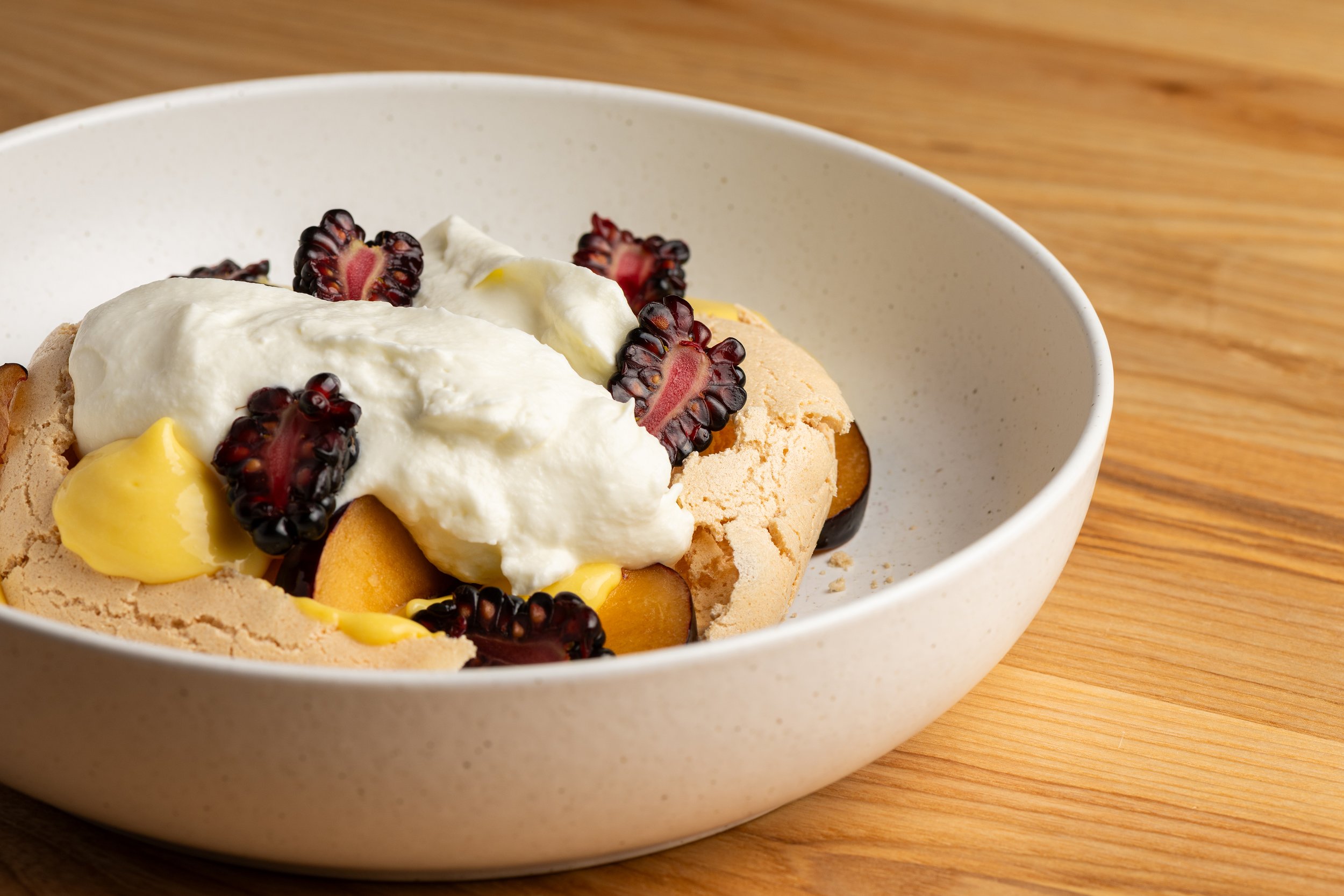 Close-up of a dessert in a white bowl with whipped cream, blackberries, and a cookie on a wooden surface.