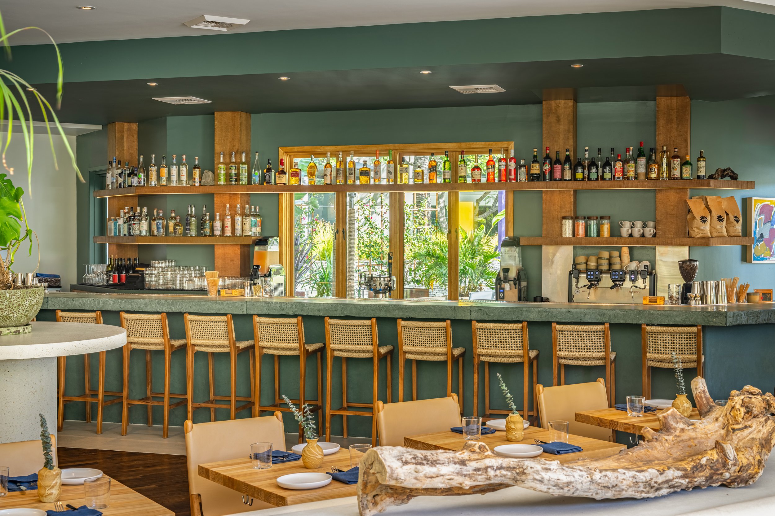 A restaurant interior with wooden seating, tables set with plates, glasses, and napkins. The focal point is a bar with bottles of alcohol on shelves behind it, and plants visible through a large window. The decor features a green painted wall and natural wooden accents.