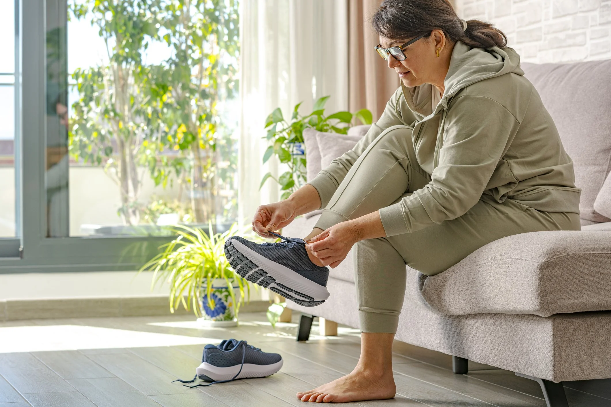 A woman sitting on a beige couch tying her shoelaces on her black and white athletic shoes. She is wearing glasses and a beige hoodie and pants. Behind her is a large window with greenery outside and a potted plant inside. The sunlight is streaming in, creating a bright atmosphere.