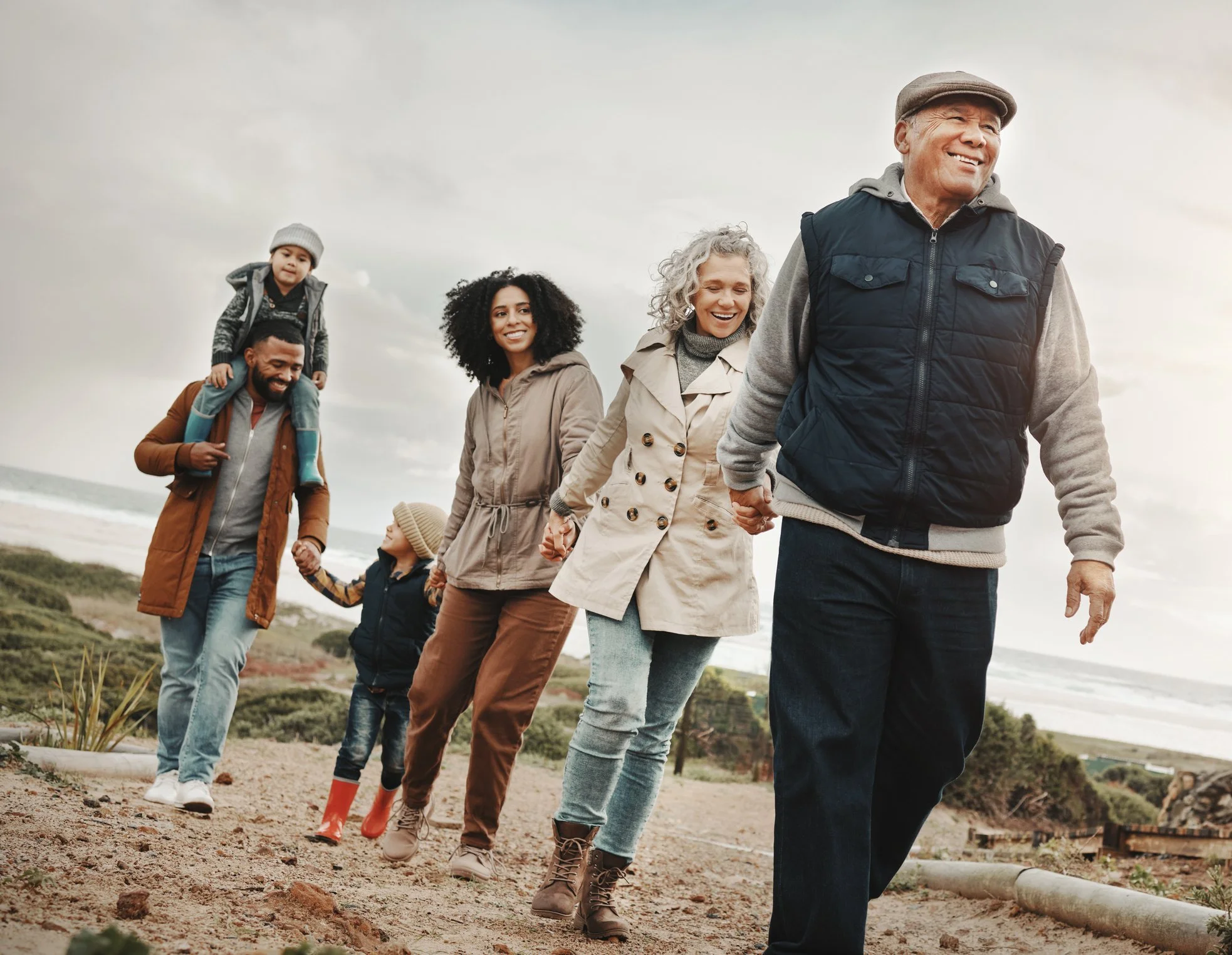 A diverse group of six people, including children and adults, walking hand in hand outdoors along a sandy trail near a beach, with cloudy skies and coastal vegetation in the background, all smiling and enjoying the moment.