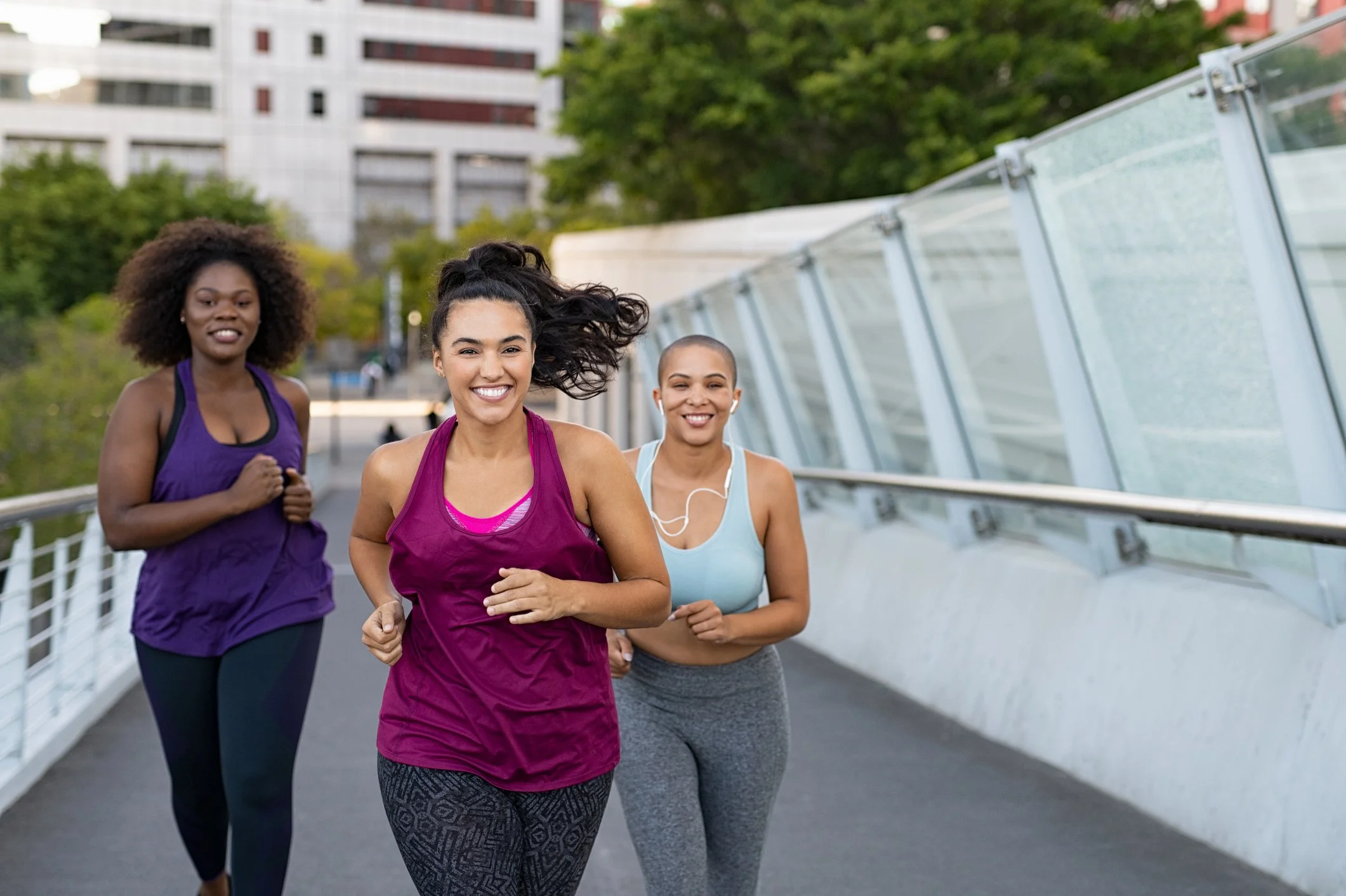 Three women running outdoors on a city path with modern buildings and green trees in the background. They are smiling and wearing athletic clothing.