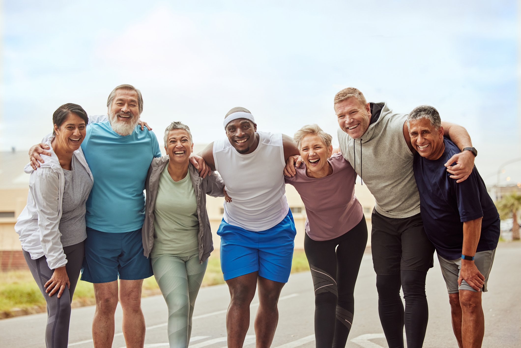 Group of seven diverse people standing close together outdoors, smiling and embracing each other after a workout or run on a sunny day.