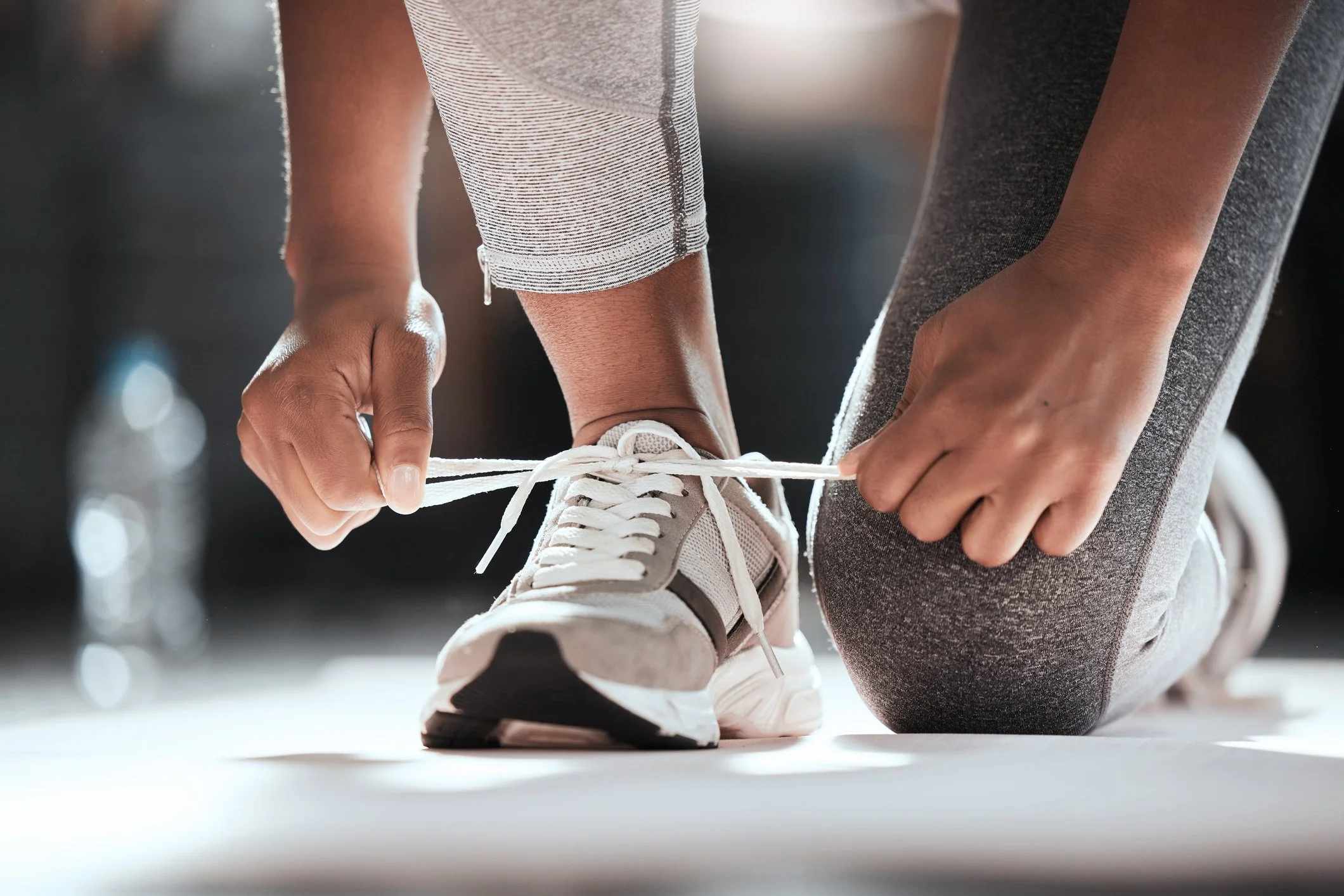 A person is tying the shoelaces of a white sneaker while kneeling on the floor, wearing gray leggings, getting prepared to be active.
