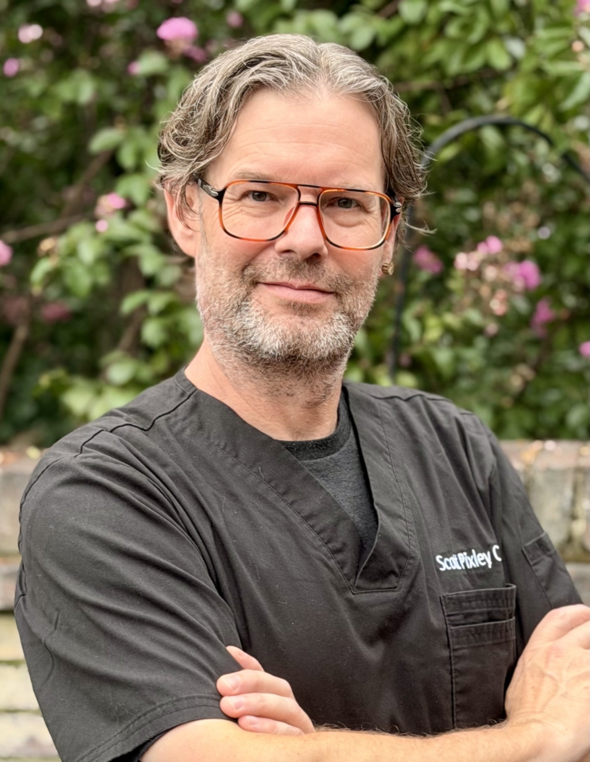 A man with brown and gray hair and beard wearing glasses and a black shirt with white text, standing outdoors with green foliage and pink flowers in the background.