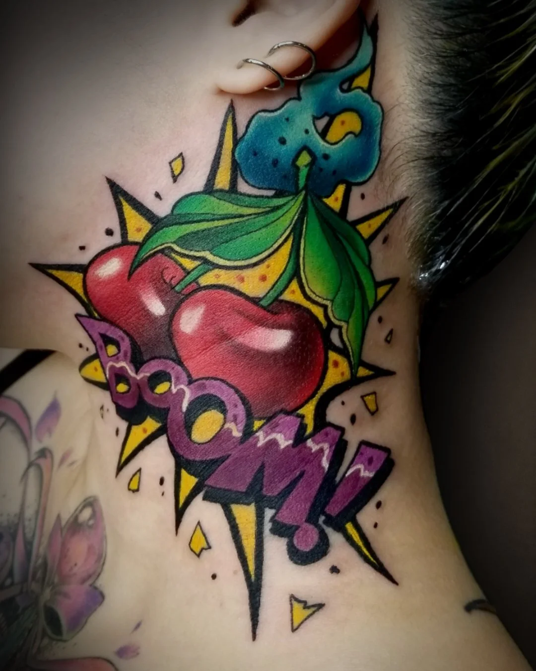 Colorful tattoo on skin featuring a pair of cherries with green leaves, a blue cloud, and the word "BOOM!" in bold letters with a yellow starburst background.