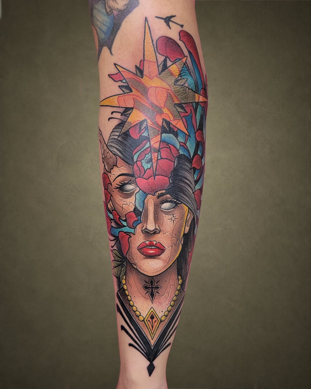 Tattoo of a woman's face with dark hair, closed eyes, red lips, and a star tattoo on her cheek, surrounded by red leaves. A large, colorful star with a lightning bolt and a black bird are above her head.