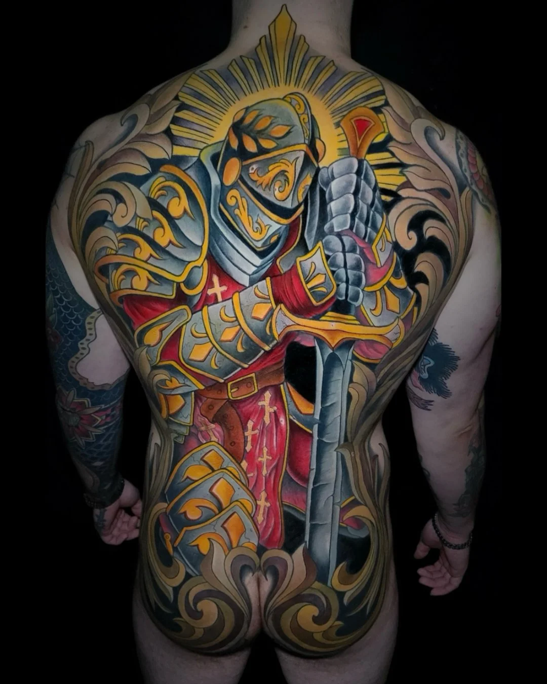Colorful back tattoo of a medieval knight in armor holding a sword, with decorative elements and a halo-like glow behind the knight's head.