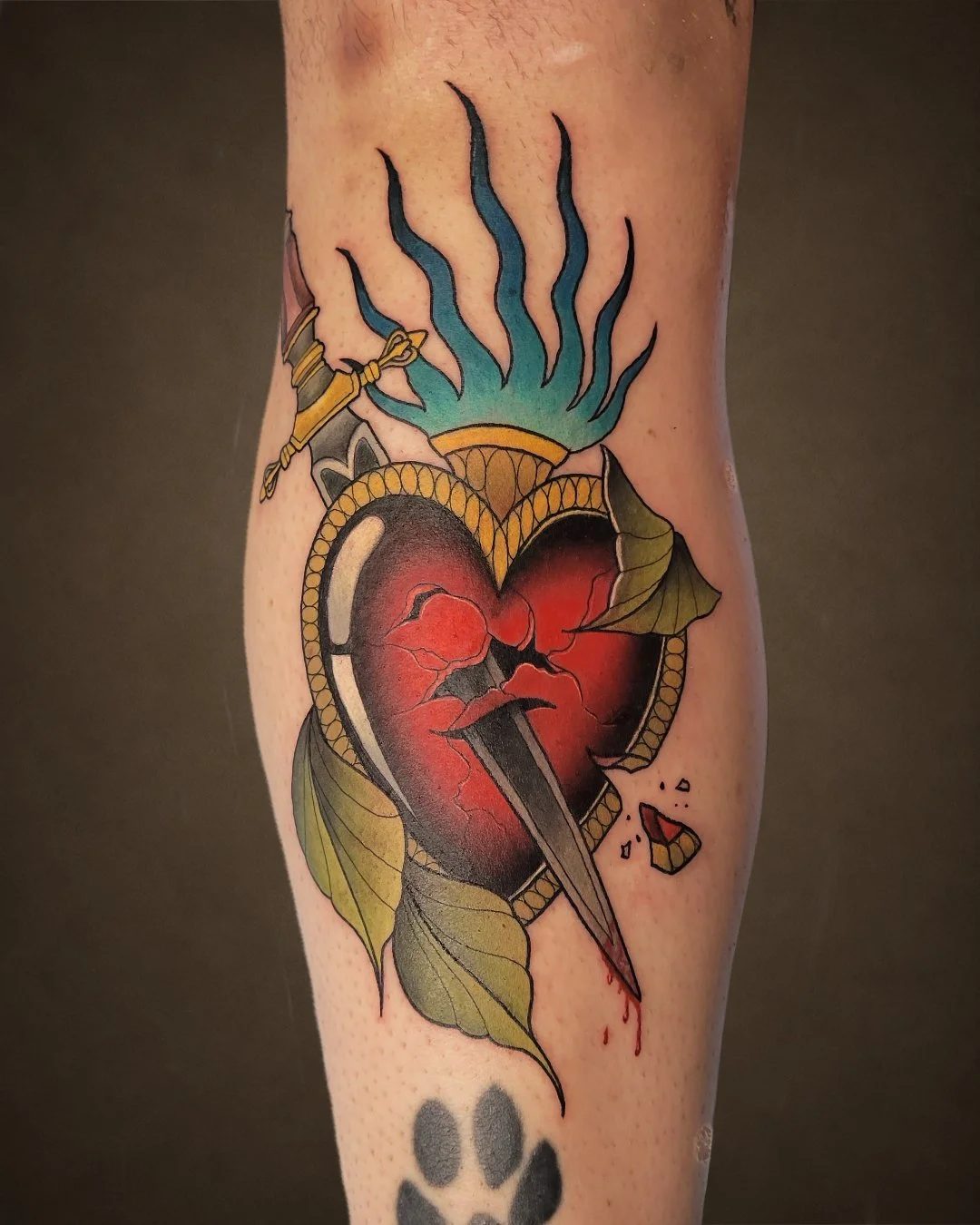 Tattoo of a cracked red heart with a dagger piercing through it, surrounded by green leaves, with blue and black flames above, and a paw print below.