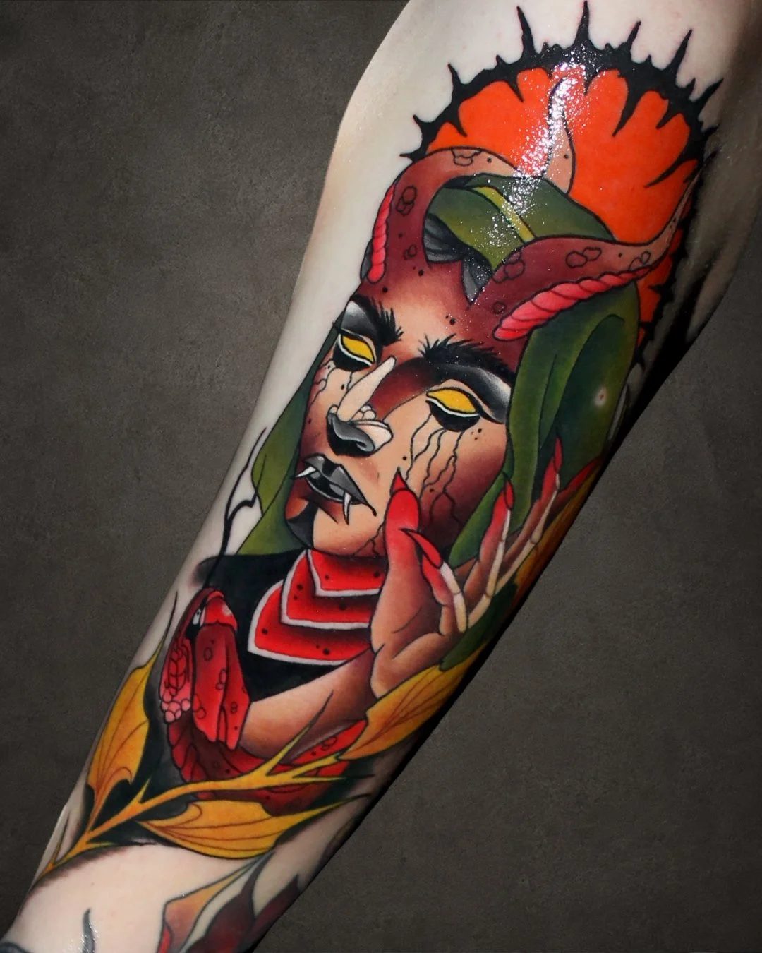 Tattoo of a woman with dark features, yellow eyes, and fangs, holding her hand near her face, surrounded by green leaves, red roses, and orange flames.
