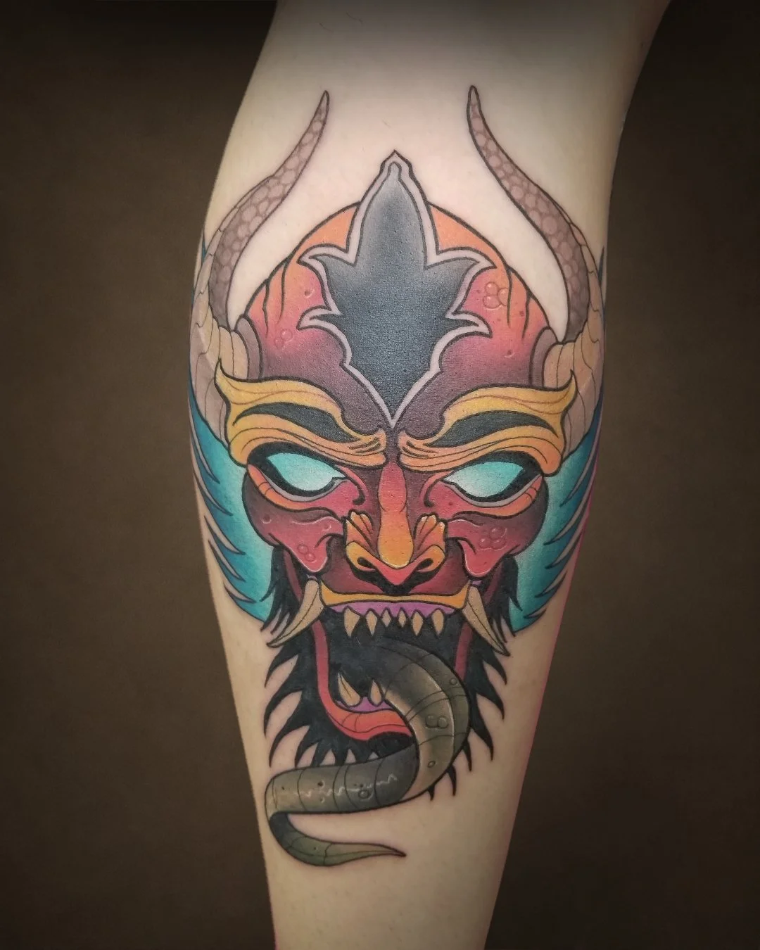 Colorful tattoo of a fierce mythical creature's face with glowing blue eyes, horns, and a serpent coming from its mouth.