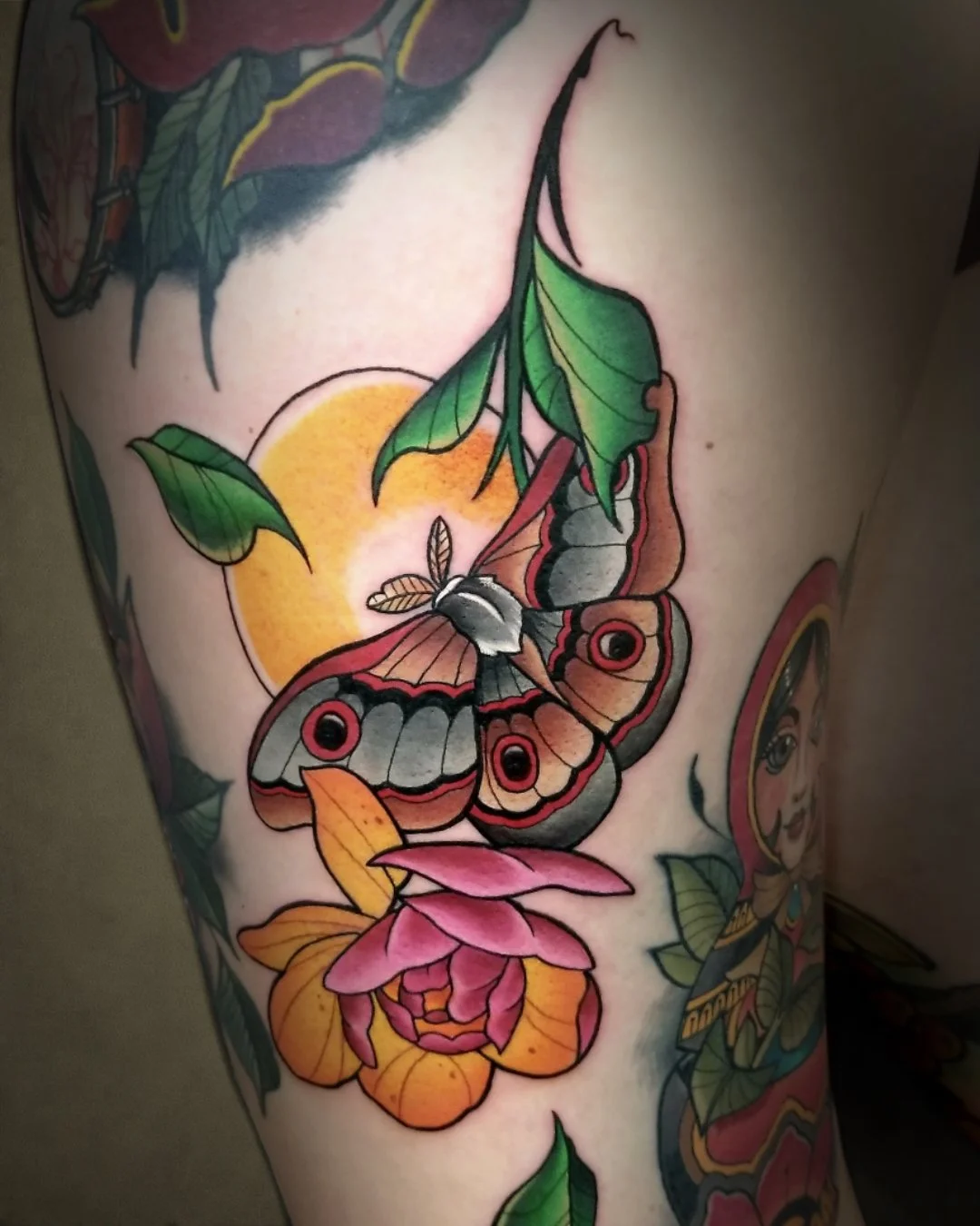 Colorful tattoo of a butterfly on a flower with a yellow moon in the background and green leaves.