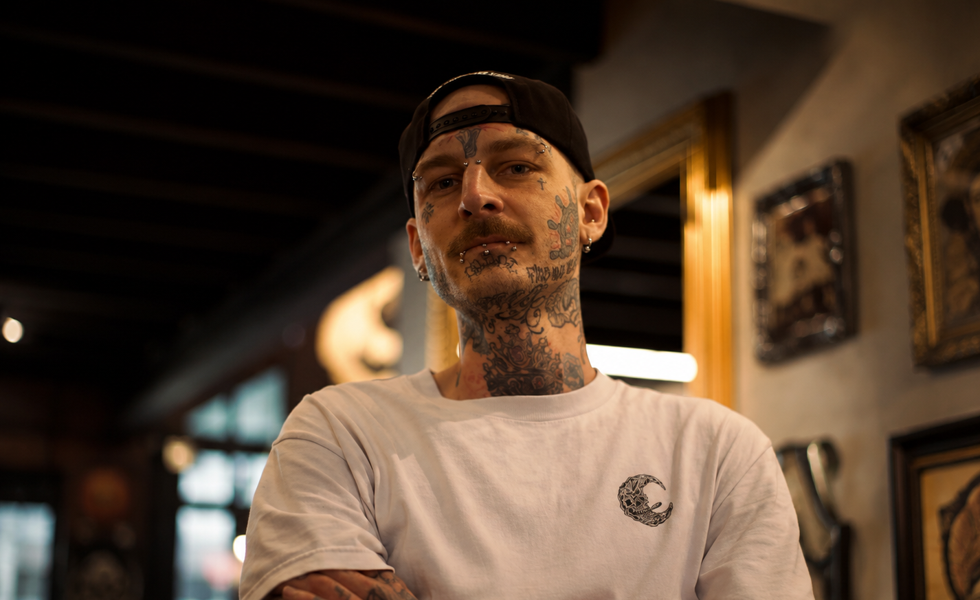 A tattooed man wearing a black cap backwards and a white T-shirt with a crescent moon logo, standing indoors with framed artwork on the wall behind him.