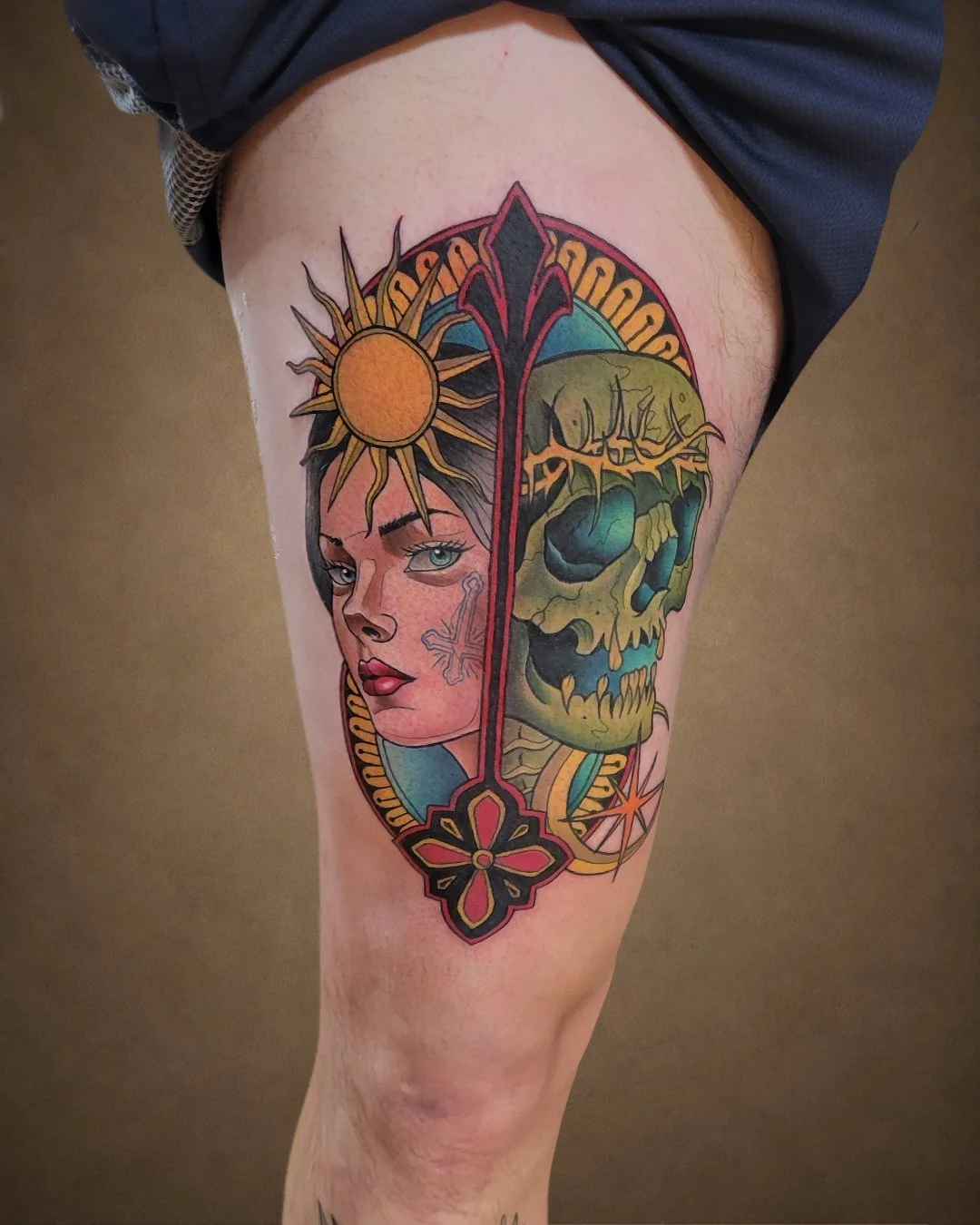 Colorful tattoo on person's thigh featuring a woman's face with a sun and a skull with a rainbow-colored backdrop.