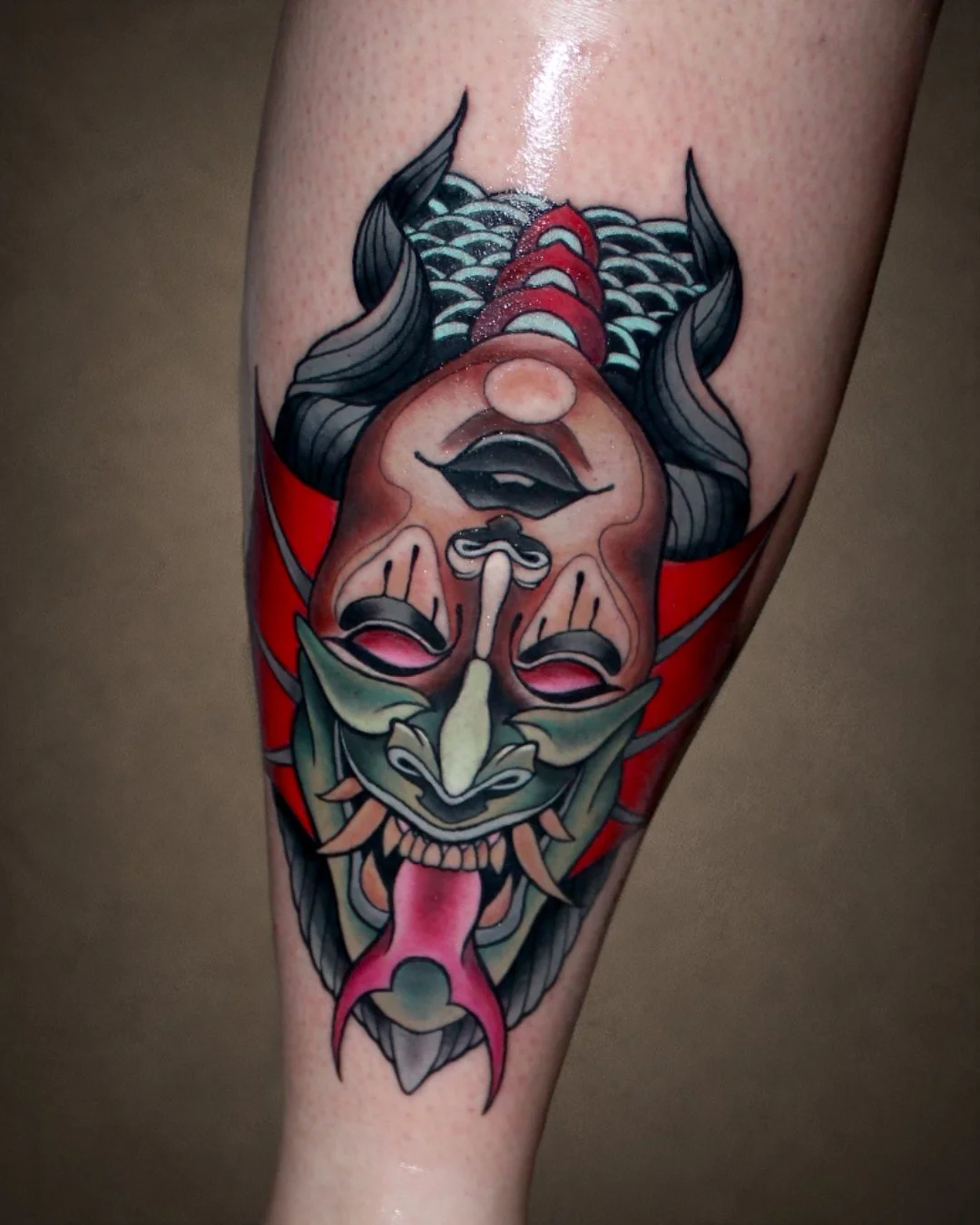 Colorful tattoo of a demonic face with horns, sharp teeth, and a long tongue, featuring intricate details and vibrant colors.
