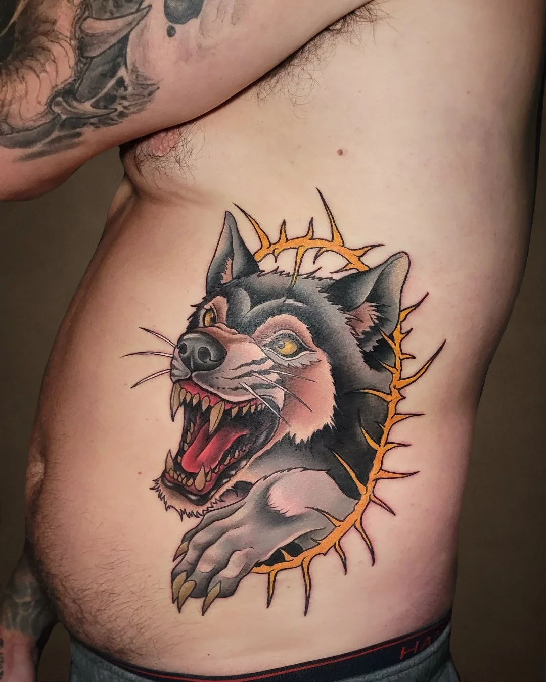 Tattoo of a snarling wolf with yellow eyes on person's side, with orange jagged lines surrounding it.