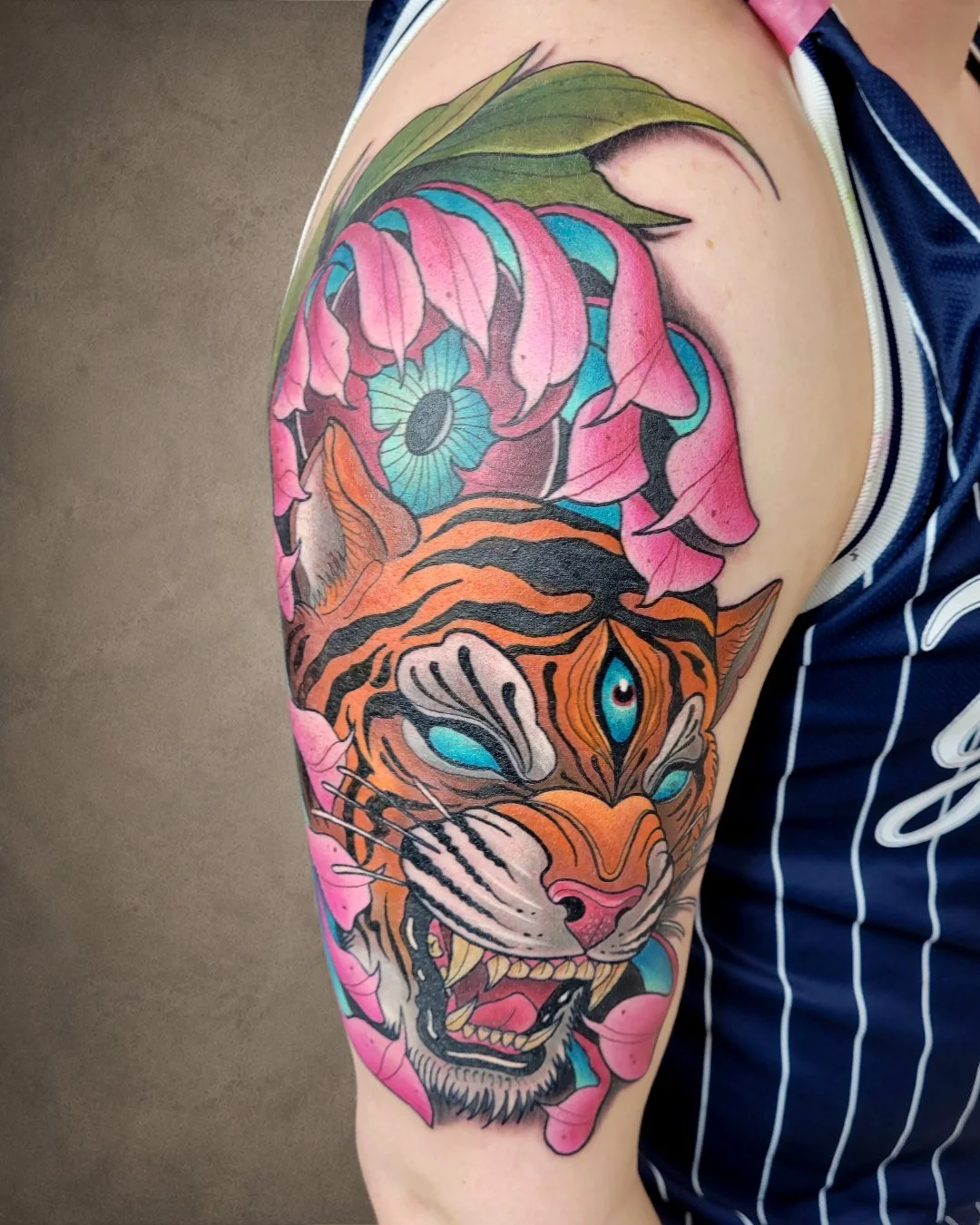 Colorful tattoo of a tiger's face with blue eyes, surrounded by pink flowers and green leaves on upper arm.