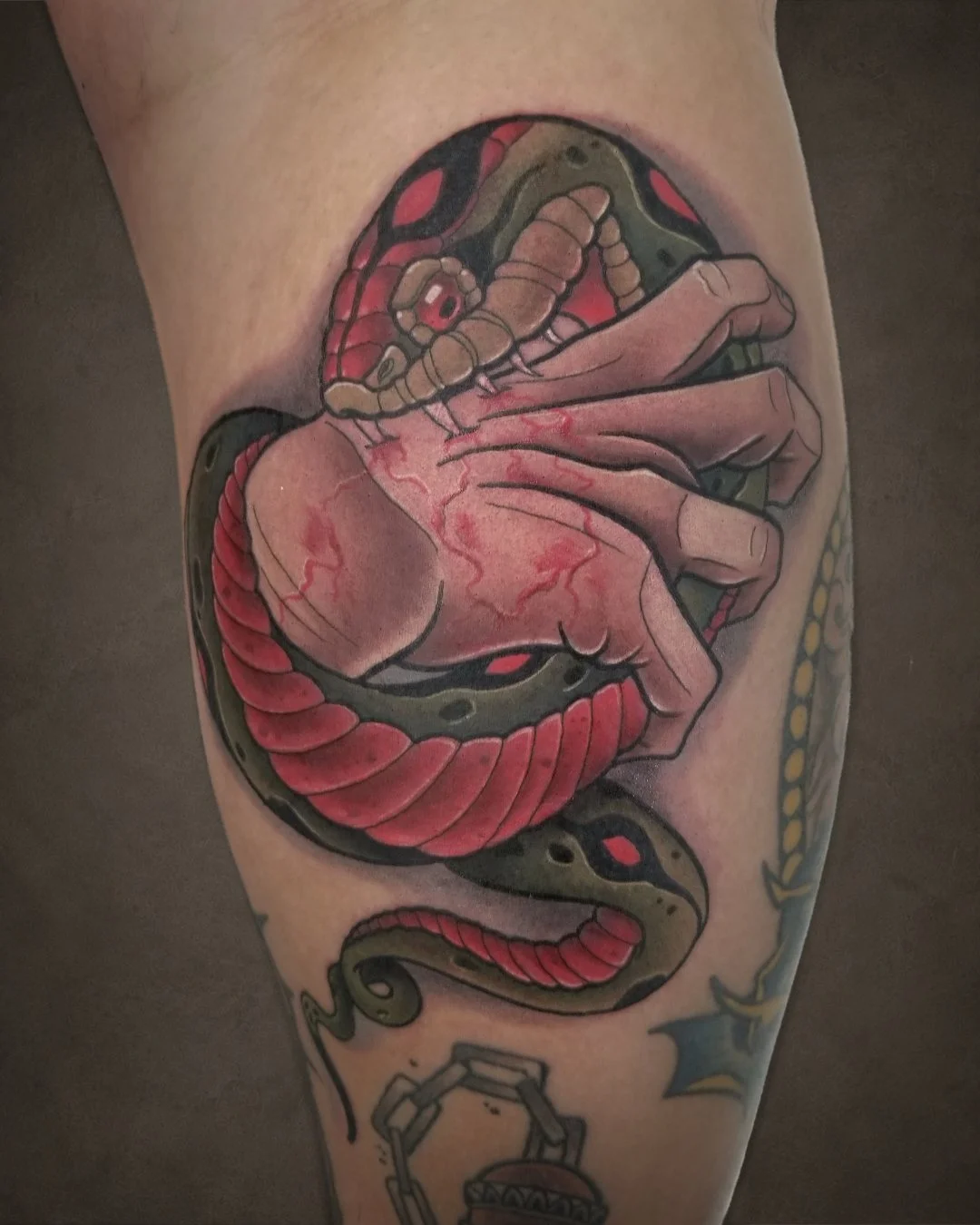 Tattoo of a realistic hand holding a human heart, surrounded by a snake. Additional tattoos include a chain with a pendant and a star with a bomb.