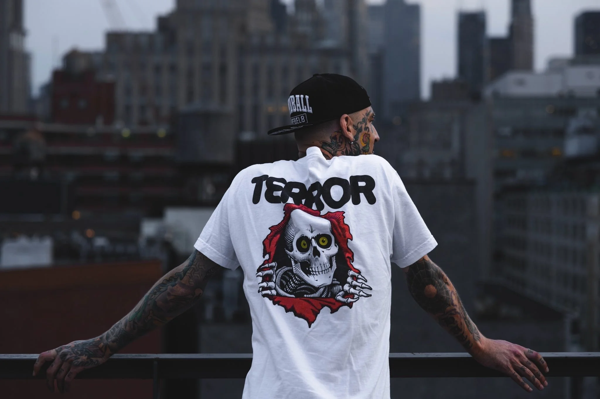A person with numerous tattoos on their arms and face, wearing a black baseball cap and a white t-shirt with a graphic of a skull and the word "TERROR" printed on the back, leaning on a railing with a cityscape of high-rise buildings in the background.
