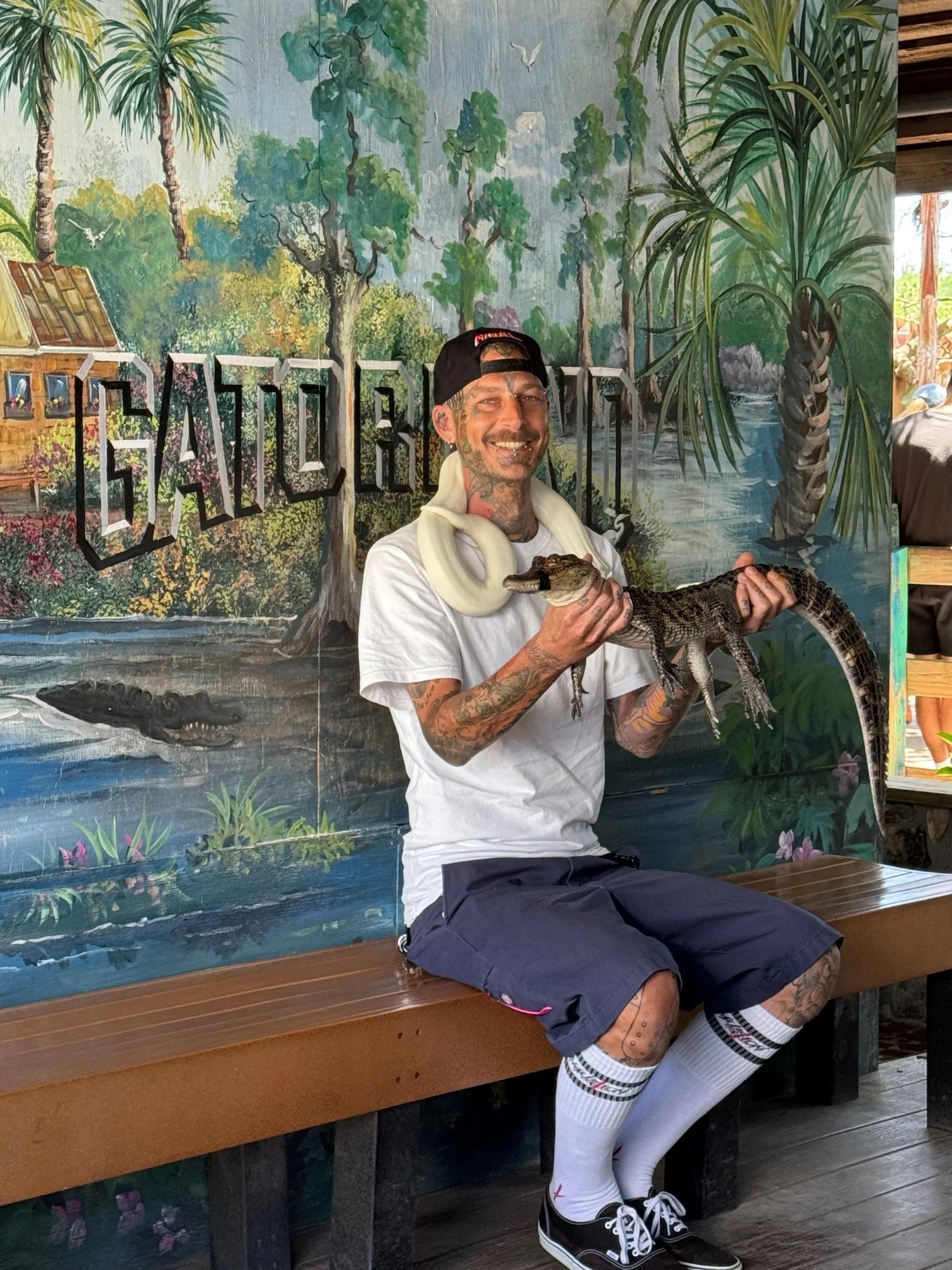 A man with tattoos, a white t-shirt, black shorts, and black sneakers sitting on a wooden bench, holding a small crocodile. He is smiling and wearing a backward black cap. Behind him, there's a colorful mural of a jungle scene with trees, water, and animals.