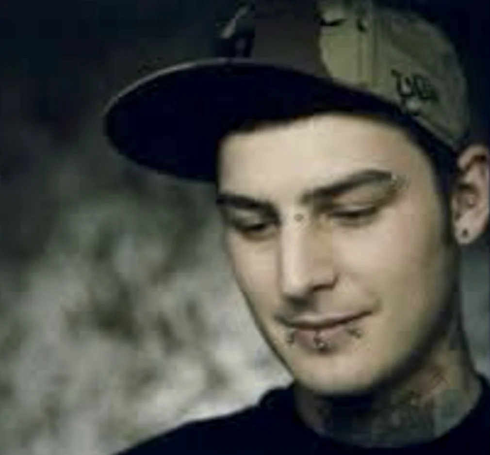 Young man with piercings in lip and ear, wearing a camo baseball cap, with closed eyes, smiling softly, smoke visible in the background.