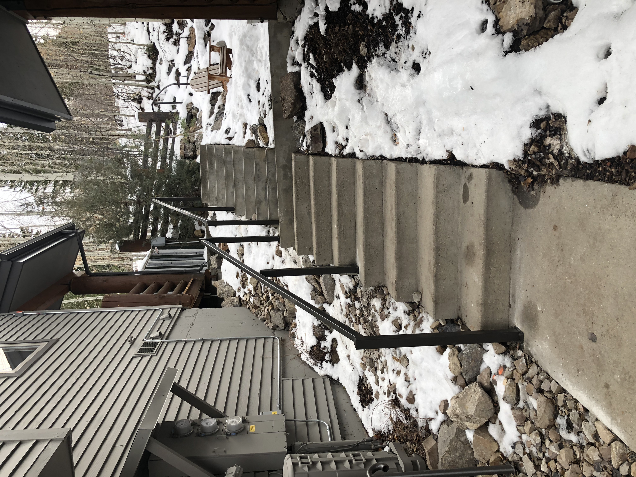 Concrete stairs with a black handrail leading up to a snow-covered yard with a wooden chair, trees, and a wooden gate in the background, adjacent to a house with beige siding and utility meters.
