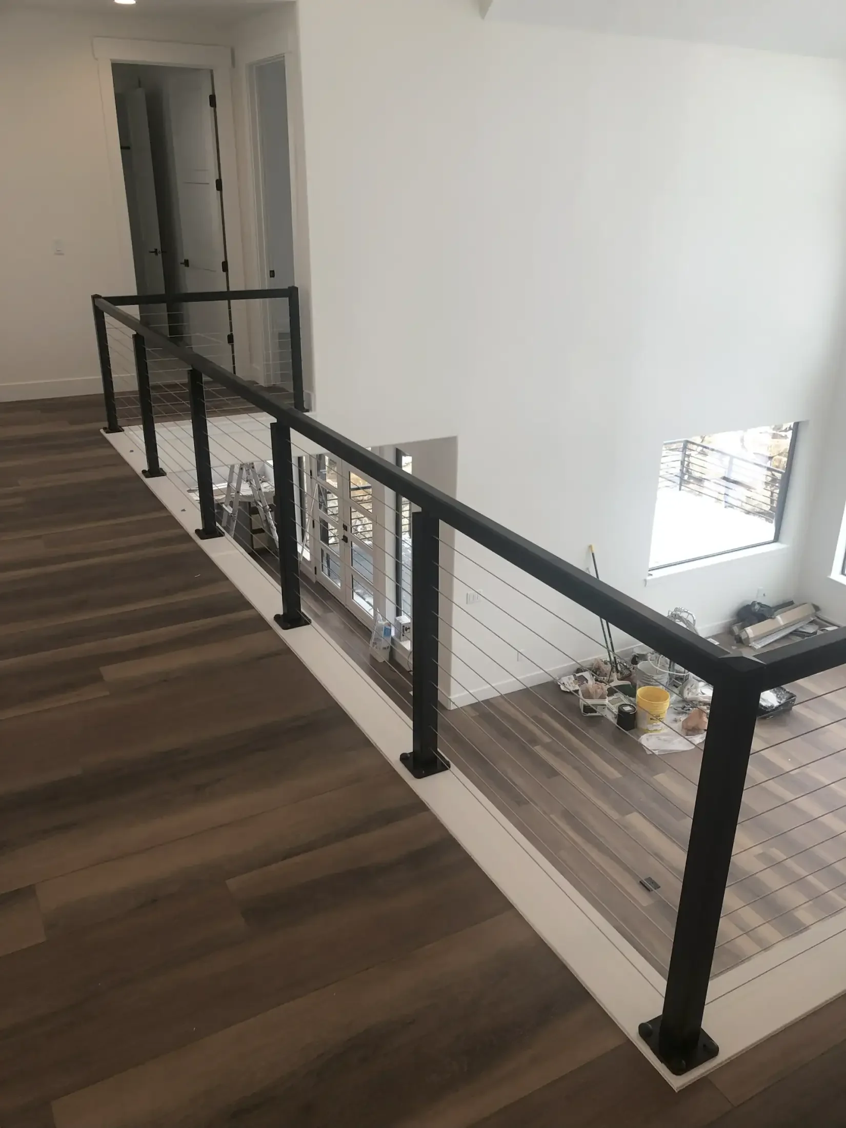 Interior view of a residential building under construction, showing a corner with a new black metal safety railing, partly installed, beside a wooden floor, and a staircase below. There are construction tools and materials on the floor, and a doorway